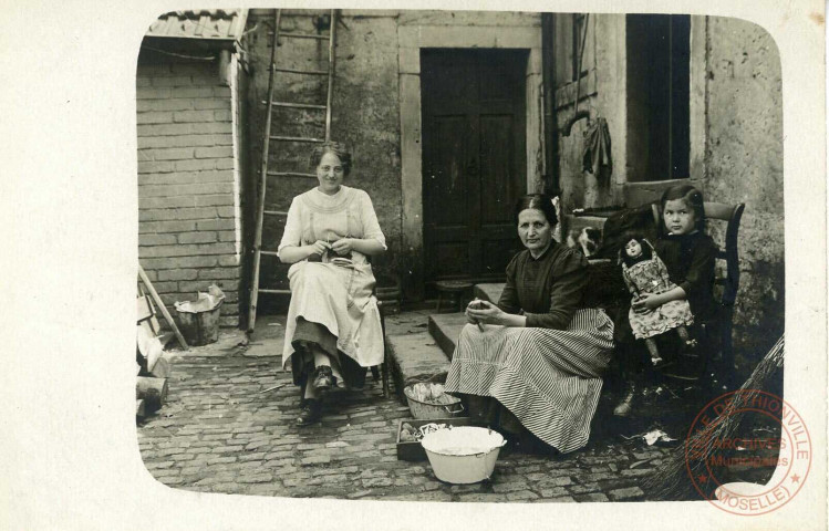[Deux femmes et une petite fille posant à l'extérieur, l'une tricote, l'autre effectue l'épluchage de pommes de terre et la petite fille tient une poupée]