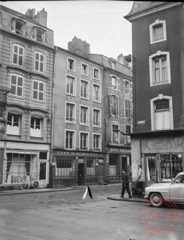 [Place au Bois. A gauche la maison du rideau, puis le café M. Schouman et un café-restaurant. Un boucher est installé dans le bâtiment à droite, on y voit des saucissons pendus dans la vitrine]