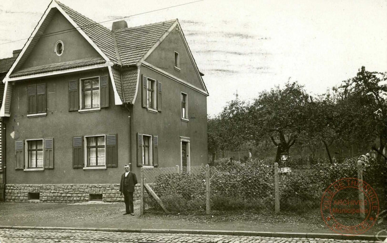 [Maison avec un homme en costume noir posant devant et un homme au fond dans le jardin]