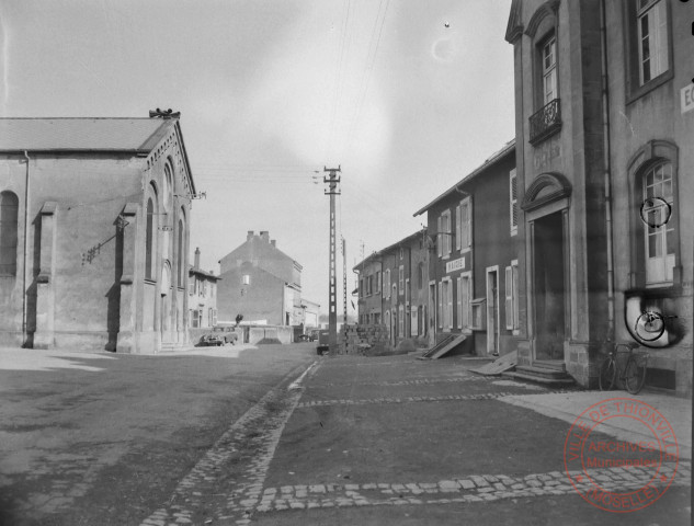 [Village du pays thionvillois non identifié. On y voit la mairie, l'école, une église et un tabac au fond]