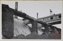 Trémies de déchargement. Photographie montrant le côté rampe inclinée des trémies de déchargement dont les poutres en béton armé sont à réparer et à munir d'une protection en traverses qui existe encore en partie. Dans le fond on aperçoit une partie des poutres en béton du bâtiment des convoyeurs centraux dont les les fers sont dénudés et fortement corrodés.