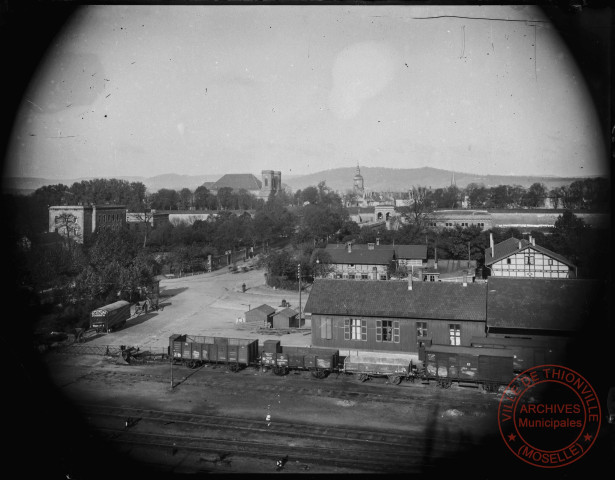 [Vue en hauteur montrant en premier plan la voie ferrée, dépôt de marchandise, non loin de la gare puis en arrière plan le pont de pierre, la porte du Pont et le centre de Thionville]