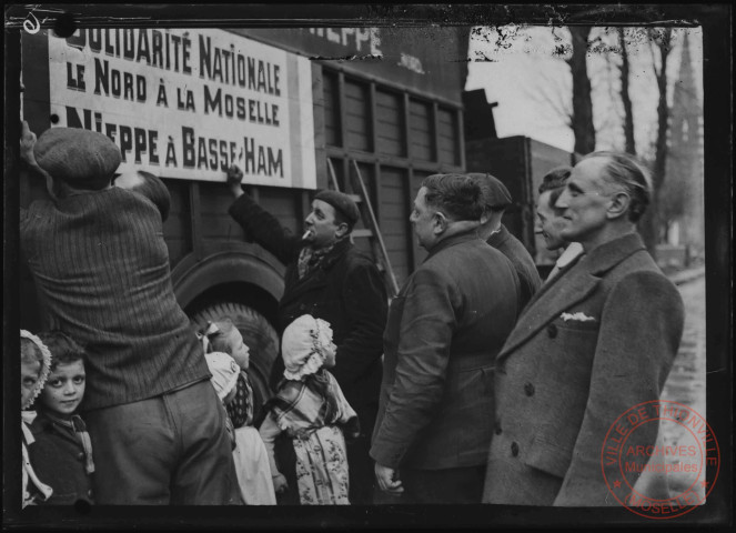 Foire exposition de 1948 - Autorités civiles et jeunes filles en costume lorrain à côté d'un camion de transports "DEREMAUX". Pancarte "Solidarité Nationale, le Nord à la Moselle, Nieppe à Basse-Ham"