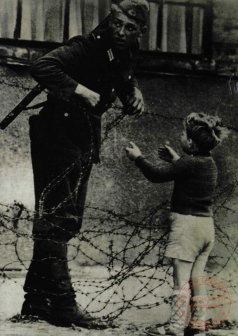 August 1961 in Berlin. Dieses Kind will hinüber. Der Posten hat Befehl, niemand durchzulassen. Trotzdem öffnet er den Draht. In diesem Augenblick sieht es der Vorgesetzte (Bild). Wegen seines Vergehens wurde der Posten sogliech abkommandiert.- Herausgebert: Verlag am Chekpoint Charlie