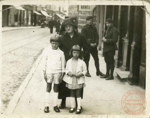 Une femme et deux enfant posent rue de Paris à Thionville. Au fond, 2 militaires