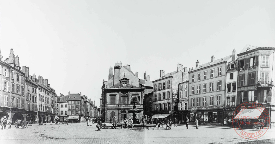 [La place du Marché avec sa fontaine au milieu, le bureau de la police juste derrière et des commerces]