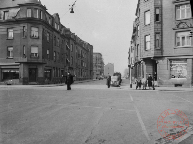 [Scène de rue à l'angle de la rue Mangin et la rue de Bourgogne. Un agent des forces de l'ordre faisant traverser la route. A gauche, boucherie-charcuterie Jung, à droite "Salon Court" articles pour écoliers]