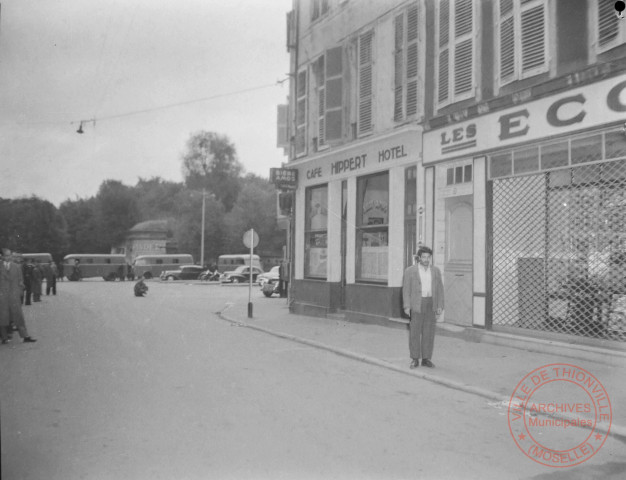 [Vue de la rue du Luxembourg, un homme à droite une arme à la main, les forces de l'ordre et leurs véhicules au fond. Dans l'angle, le café et hôtel Hippert. Il semblerait que ce soit une reconstitution d'une affaire criminelle]