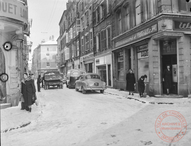 [Rue Jemmapes sous la neige. Plusieurs personnes déneigent la route. On peut voir les commerces suivants, Chaussures Weiler, Romu, Henri Daster]