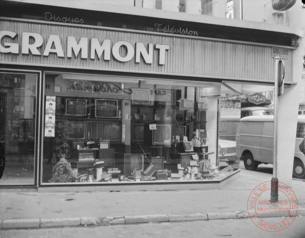 [Magasin Zosi, disques et télévision (Grammont marque de télévision), à l'angle de la place au Bois et la rue du Four Banal, en face (se reflétant dans la vitrine), le café Schouman au numéro 19 rue du Four Banal]