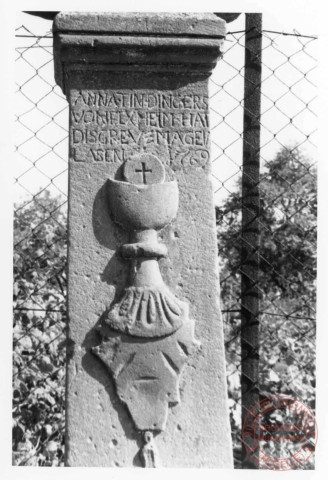 Bildstock situé à l'angle de la rue de Trèves et la route de Rodemack à Fixem. Croix érigée en 1769 (date portée sur le fût) pour Anne Tindingers. Portement de croix, calice, fleur de lys, feuillage, fleur