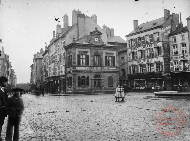 [La place du Marché et la rue du Luxembourg. Au fond la porte du Luxembourg. Au premier plan la police municipale, photographie Bruère, Au Bon Marché, magasin de confection de vêtements Marx-Weil, pharmacie]